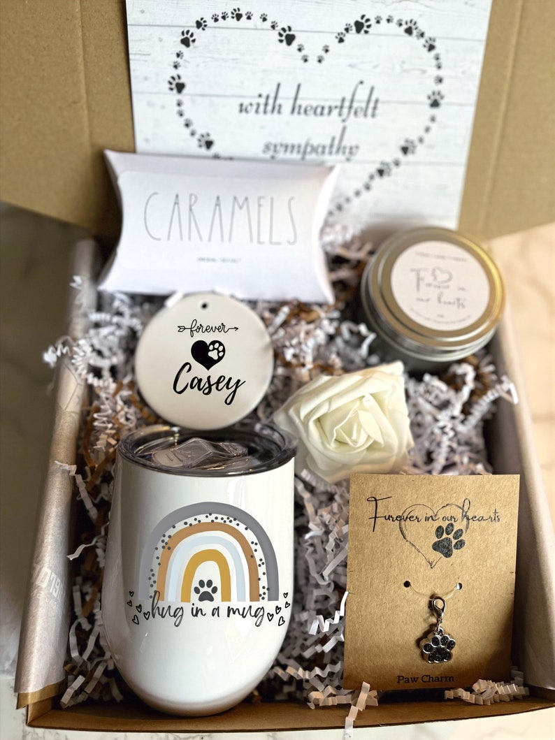 May include: A sympathy gift box with a white ceramic ornament that says "forever Casey", a white ceramic mug with a rainbow and paw print design that says "hug in a mug", a small white box with the word "caramels" on it, a candle with a paw print and the words "Forever in our hearts", and a silver paw print charm on a brown card that says "Forever in our hearts" and "Paw Charm".
