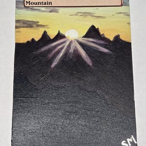 May include: A rectangular trading card featuring a hand-painted landscape of dark, jagged mountains silhouetted against a yellow and orange sunset. The word Mountain is printed at the top. A bright sun emits light rays over the peaks. The initials SM are in the corner.