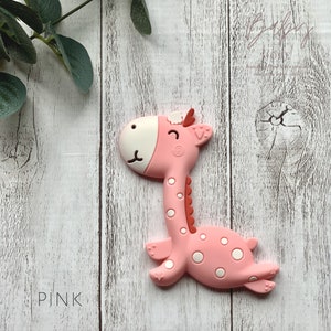 May include: Pink silicone giraffe teether with white spots and a smiling face. The teether is on a white wooden background.