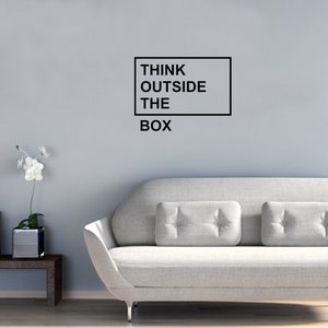 May include: A gray couch with two pillows in a living room with a white wall. The wall has a black framed sign that reads "Think Outside The Box".