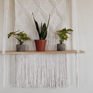 May include: A white macrame wall hanging with a wooden shelf displaying three potted plants. The plants include a snake plant and two others with green leaves. The macrame has a diamond pattern and fringe.