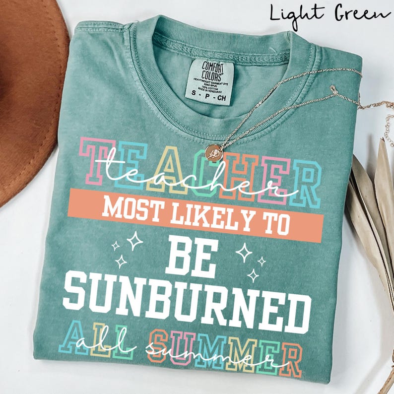 May include: A light green t-shirt with the text "Teacher Most Likely To Be Sunburned All Summer" in colorful block letters. The shirt has a crew neck and short sleeves. A silver necklace with a pendant is draped on the shirt.