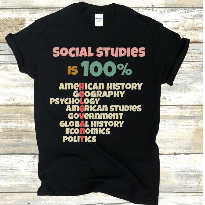 Op de afbeelding: Zwart T-shirt met de tekst "SOCIAL STUDIES IS 100%" en een lijst met vakken, waaronder Amerikaanse geschiedenis, geografie, psychologie, Amerikaanse studies, overheid, wereldgeschiedenis, economie en politiek.