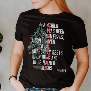 May include: Black t-shirt with a Christmas tree design featuring the words "A Child Has Been Born For Us, A Son Is Given To Us, Authority Rests Upon Him: And He Is Named Jesus" and the scripture reference "Isaiah 9:6".