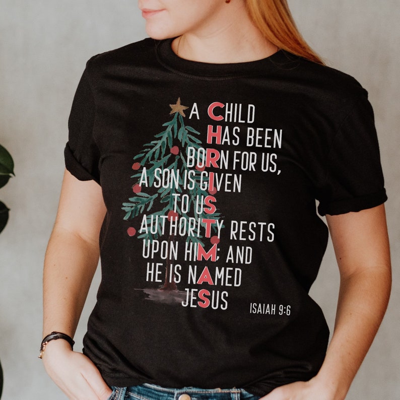 May include: Black t-shirt with a Christmas tree design featuring the words "A Child Has Been Born For Us, A Son Is Given To Us, Authority Rests Upon Him: And He Is Named Jesus" and the scripture reference "Isaiah 9:6".