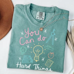 Peut inclure: Un t-shirt vert sarcelle avec le texte "You Can Do Hard Things" en rose et jaune. Le t-shirt présente une ampoule, un globe terrestre, une règle, une boussole, une pomme et un carnet.