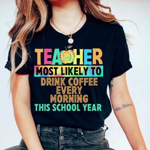 以下が含まれることがあります： 「Teacher most likely to drink coffee every morning this school year」というテキストが書かれた、カラフルなタイダイ柄の黒いTシャツです。
