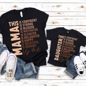 May include: Two black t-shirts with the text "This is confident is strong is blessed is motivated is elevated is melanated is happy is beautiful is enough is me" and "This is confident is strong is blessed is motivated is elevated is melanated is happy is beautiful is enough is" on them. The larger shirt says "Mama" and the smaller shirt says "Daughter".