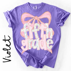 May include: A violet short-sleeve t-shirt featuring a large pink bow graphic with gold glitter outlines. The text fifth grade is printed in the center, with letters filled by a light pink gingham pattern and a blue and white floral print design.
