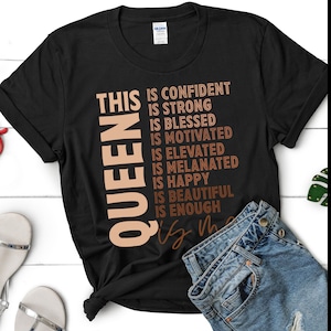 May include: Black t-shirt with the text "THIS QUEEN IS CONFIDENT IS STRONG IS BLESSED IS MOTIVATED IS ELEVATED IS MELANATED IS HAPPY IS BEAUTIFUL IS ENOUGH" in brown and orange lettering.