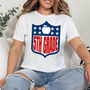 May include: White t-shirt with a blue and red graphic of a football helmet with a red apple and the text "5th Grade".