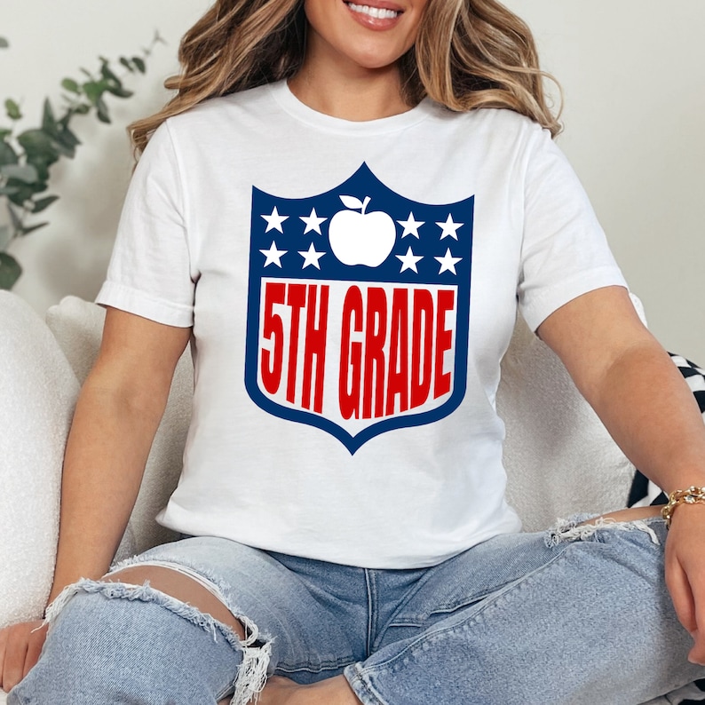 May include: White t-shirt with a blue and red graphic of a football helmet with a red apple and the text "5th Grade".