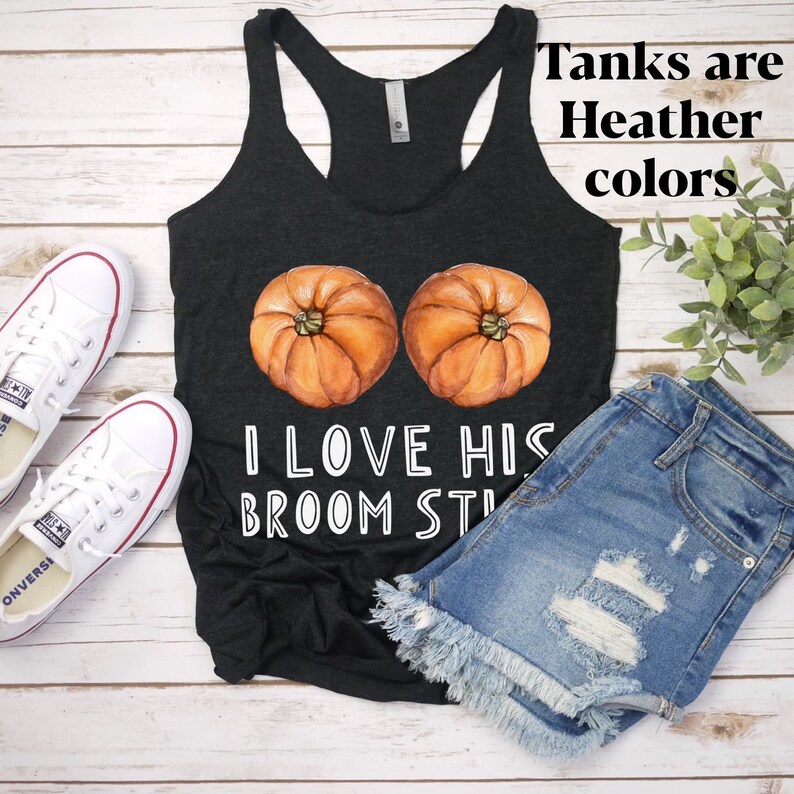 May include: Dark gray racerback tank top with a watercolor design of two pumpkins and the text "I LOVE HIS BROOM STICK". The text "Tanks are Heather colors" is also visible. The tank top is paired with denim shorts and white sneakers.