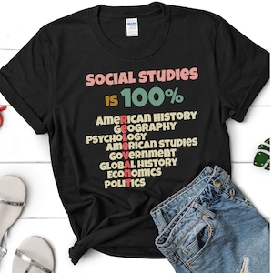 Op de afbeelding: Zwart T-shirt met de tekst "SOCIAL STUDIES IS 100%" en een lijst met vakken, waaronder Amerikaanse geschiedenis, geografie, psychologie, Amerikaanse studies, overheid, wereldgeschiedenis, economie en politiek.