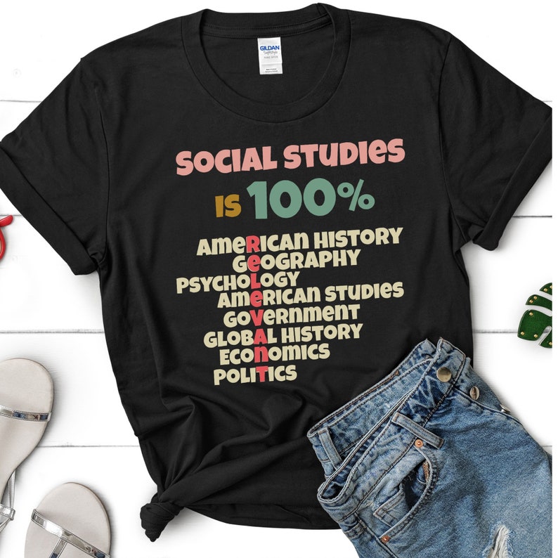 Op de afbeelding: Zwart T-shirt met de tekst "SOCIAL STUDIES IS 100%" en een lijst met vakken, waaronder Amerikaanse geschiedenis, geografie, psychologie, Amerikaanse studies, overheid, wereldgeschiedenis, economie en politiek.