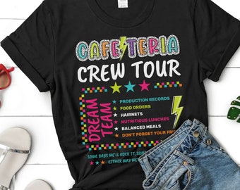 May include: Black t-shirt with the text "Cafeteria Crew Tour" in colorful letters. The text "Dream Team" is in a colorful checkered pattern with stars and a lightning bolt. The text also includes a list of tasks: "Production Records", "Food Orders", "Hairnets", "Nutritious Lunches", "Balanced Meals", and "Don't Forget Your Fri". The text "Some days we'll rock it, some days we won't. Either way we'll have fun!" is at the bottom.