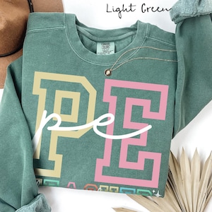 May include: Light green sweatshirt with the letters "PE" in a block letter style, with the word "pe" in cursive. The "P" is tan, and the "E" is pink. The text "Light Green" is at the top. A gold necklace is visible.