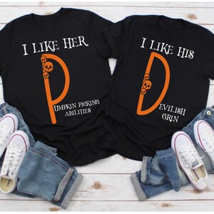 May include: Two black t-shirts with orange lettering and skull graphics. The text on the shirts reads "I like her" and "I like his". The shirts are laid out on a wooden surface with blue jeans and white sneakers.