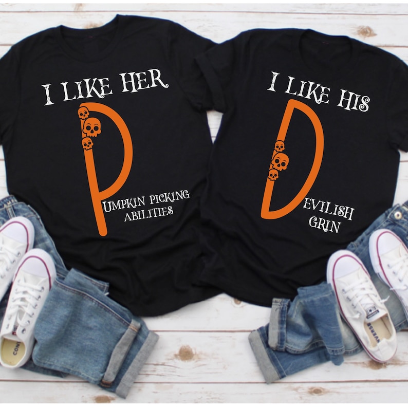 May include: Two black t-shirts with orange lettering and skull graphics. The text on the shirts reads "I like her" and "I like his". The shirts are laid out on a wooden surface with blue jeans and white sneakers.