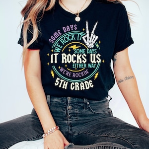 May include: Black t-shirt with a colorful graphic design that says "Some days we rock it, some days it rocks us, either way we're rockin' 5th grade". The graphic includes a skeleton hand making a peace sign.