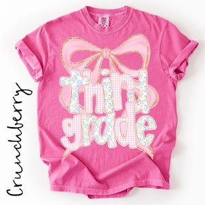 May include: A bright pink short-sleeved t-shirt featuring a large graphic of a pink bow with gold glitter outlines. The text "third grade" is printed in a mix of light pink gingham and blue floral patterns, centered on the front of the shirt.
