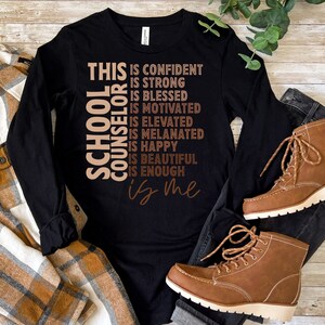 May include: Black long-sleeve shirt with the text "THIS IS CONFIDENT, IS STRONG, IS BLESSED, IS MOTIVATED, IS ELEVATED, IS MELANATED, IS HAPPY, IS BEAUTIFUL, IS ENOUGH, is me" and "SCHOOL COUNSELOR" in a vertical design. Brown boots and a plaid shirt are also visible.