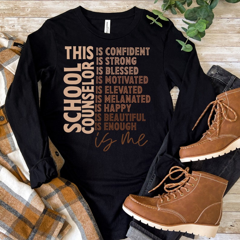 May include: Black long-sleeve shirt with the text "THIS IS CONFIDENT, IS STRONG, IS BLESSED, IS MOTIVATED, IS ELEVATED, IS MELANATED, IS HAPPY, IS BEAUTIFUL, IS ENOUGH, is me" and "SCHOOL COUNSELOR" in a vertical design. Brown boots and a plaid shirt are also visible.