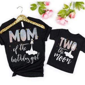 Pode incluir: Duas camisetas pretas com tema espacial. A camiseta maior diz "MOM of the birthday girl" com um foguete e estrelas. A camiseta menor diz "TWO the moon" com um foguete e estrelas.