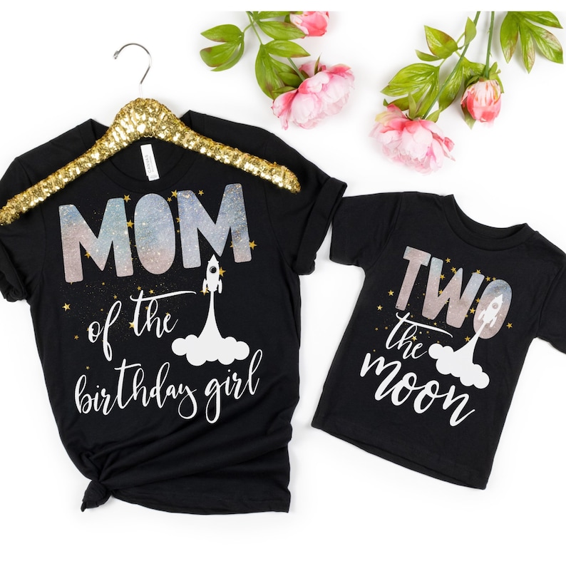 Pode incluir: Duas camisetas pretas com tema espacial. A camiseta maior diz "MOM of the birthday girl" com um foguete e estrelas. A camiseta menor diz "TWO the moon" com um foguete e estrelas.