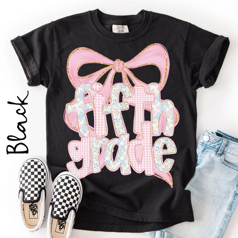 May include: A black short-sleeved t-shirt featuring a large graphic of a pink bow with gold glitter edges. The text "fifth grade" is printed in a bubbly font with a pink gingham and blue floral pattern, layered over the bow design.
