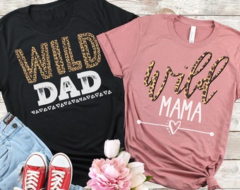 Puede incluir: Dos camisetas con un diseño de estampado de leopardo. La camiseta negra dice "Wild Dad" en letras blancas con un patrón tribal debajo. La camiseta rosa dice "Wild Mama" en letras blancas con un pequeño corazón debajo.