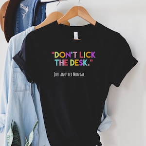 K&ouml;nnte beinhalten: Schwarzes T-Shirt mit dem Text "DON'T LICK THE DESK." in bunten Buchstaben und dem Text "JUST ANOTHER MONDAY." in wei&szlig;en Buchstaben.
