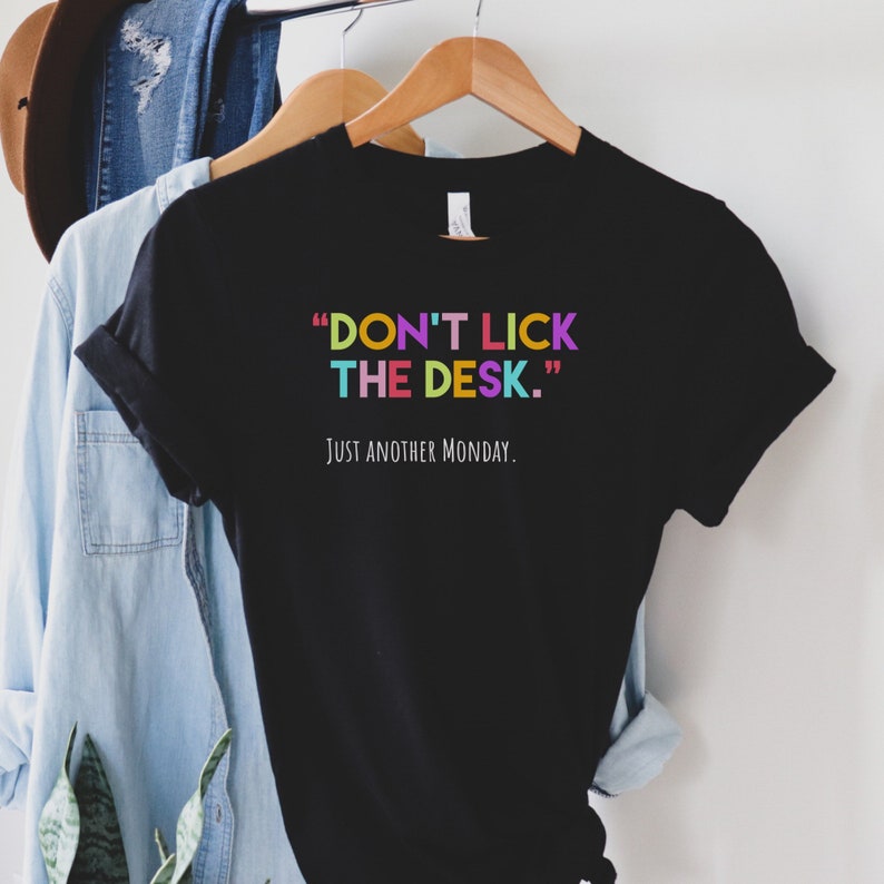 K&ouml;nnte beinhalten: Schwarzes T-Shirt mit dem Text "DON'T LICK THE DESK." in bunten Buchstaben und dem Text "JUST ANOTHER MONDAY." in wei&szlig;en Buchstaben.