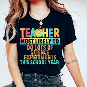 以下が含まれることがあります： タイダイ柄の黒いTシャツに「TEACHER MOST LIKELY TO DO LOTS OF SCIENCE EXPERIMENTS THIS SCHOOL YEAR」という文字が書かれています。