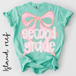 May include: A mint green short-sleeve t-shirt featuring a large pink bow graphic with gold glitter outlines. The text "second grade" is printed in a mix of pink gingham and blue floral patterns, centered over the bow on the front of the shirt.