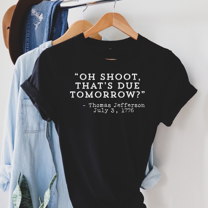 Op de afbeelding: Zwart t-shirt met de quote "OH SHOOT, THAT'S DUE TOMORROW?" toegeschreven aan Thomas Jefferson, 3 juli 1776, in witte tekst. Het t-shirt hangt aan een houten hanger, met andere kledingstukken op de achtergrond.