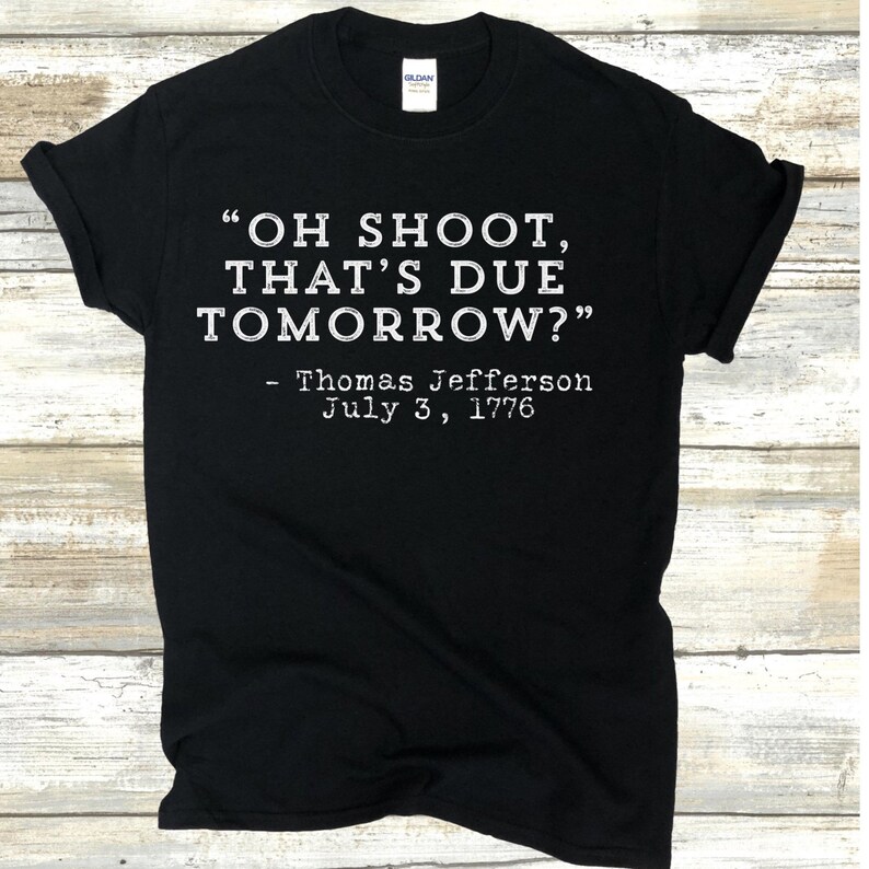 Op de afbeelding: Zwart T-shirt met witte tekst. De tekst luidt "OH SHOOT, THAT'S DUE TOMORROW?" gevolgd door "- Thomas Jefferson July 3, 1776." Het shirt is van het merk Gildan.