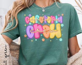 Puede incluir: Camiseta verde claro con las palabras "Cafeteria Crew" en letras coloridas. El diseño incluye flores y estrellas. La camiseta es de cuello redondo y tiene mangas cortas.