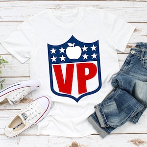 May include: White t-shirt with a red and blue shield design. The shield has a white apple with a stem and leaf inside and the letters "VP" in red. The shield is surrounded by white stars on a blue background.