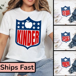 May include: White t-shirt with a blue and red shield design featuring a white apple and the word "kinder" in red letters. The shield has white stars on a blue background. The t-shirt is being modeled by a person wearing blue jeans.