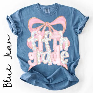 May include: A blue denim-colored short-sleeve t-shirt featuring a large pink bow graphic above the text "fifth grade". The lettering is filled with a pattern of pink gingham and small blue floral designs, outlined in a glittery gold-toned border.