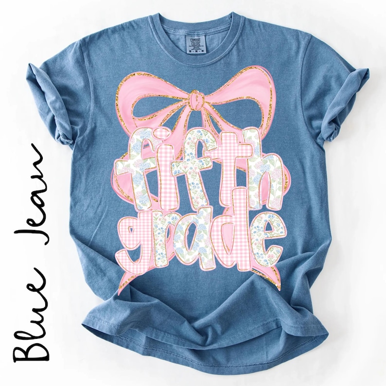 May include: A blue denim-colored short-sleeve t-shirt featuring a large pink bow graphic above the text "fifth grade". The lettering is filled with a pattern of pink gingham and small blue floral designs, outlined in a glittery gold-toned border.