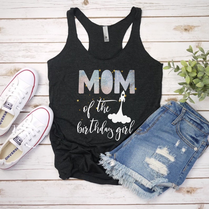 Pode incluir: Camisola de al&ccedil;as preta com um gr&aacute;fico de tem&aacute;tica gal&aacute;ctica que diz "MOM of the birthday girl" com um foguete e estrelas.