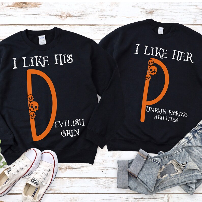 May include: Two black sweatshirts with orange text and graphics. One sweatshirt says "I like his D Evilish Grin" with a skull and crossbones graphic. The other sweatshirt says "I like her D Umpkin Picking Abilities" with a skull and crossbones graphic.
