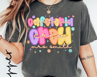 Puede incluir: Camiseta gris oscuro con las palabras "Cafeteria Crew Mrs. Smith" en letras coloridas. El diseño incluye flores y estrellas. La camiseta es de estilo casual.