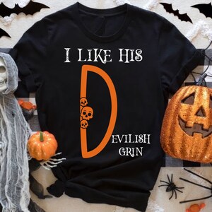 May include: Black t-shirt with the text "I like his D EVILISH GRIN" in white. The letter D is orange and has three orange skulls behind it.