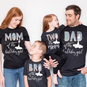 Pode incluir: Um conjunto de quatro camisetas pretas com um design tem&aacute;tico espacial. As camisetas apresentam o texto "M&atilde;e da menina do anivers&aacute;rio", "Irm&atilde;o da menina do anivers&aacute;rio", "Dois a lua" e "Pai da menina do anivers&aacute;rio".