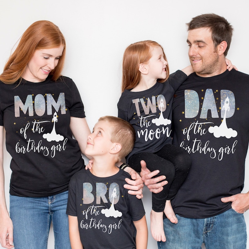 Pode incluir: Um conjunto de quatro camisetas pretas com um design tem&aacute;tico espacial. As camisetas apresentam o texto "M&atilde;e da menina do anivers&aacute;rio", "Irm&atilde;o da menina do anivers&aacute;rio", "Dois a lua" e "Pai da menina do anivers&aacute;rio".