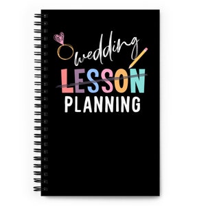 May include: A black spiral notebook with the text "Wedding Lesson Planning" in colorful letters. The word "Lesson" is crossed out with a pencil and a ring with a heart is above the word "Wedding".