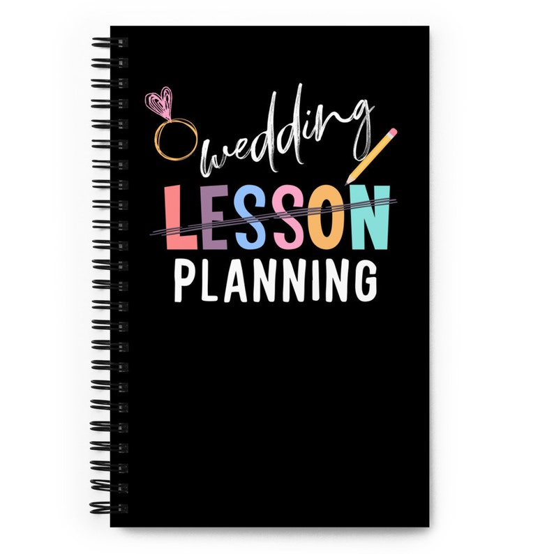 May include: A black spiral notebook with the text "Wedding Lesson Planning" in colorful letters. The word "Lesson" is crossed out with a pencil and a ring with a heart is above the word "Wedding".